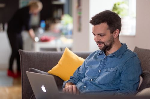 Ung mann med skjegg og jeansskjorte sitter smilende i sofaen, med en laptop på fanget