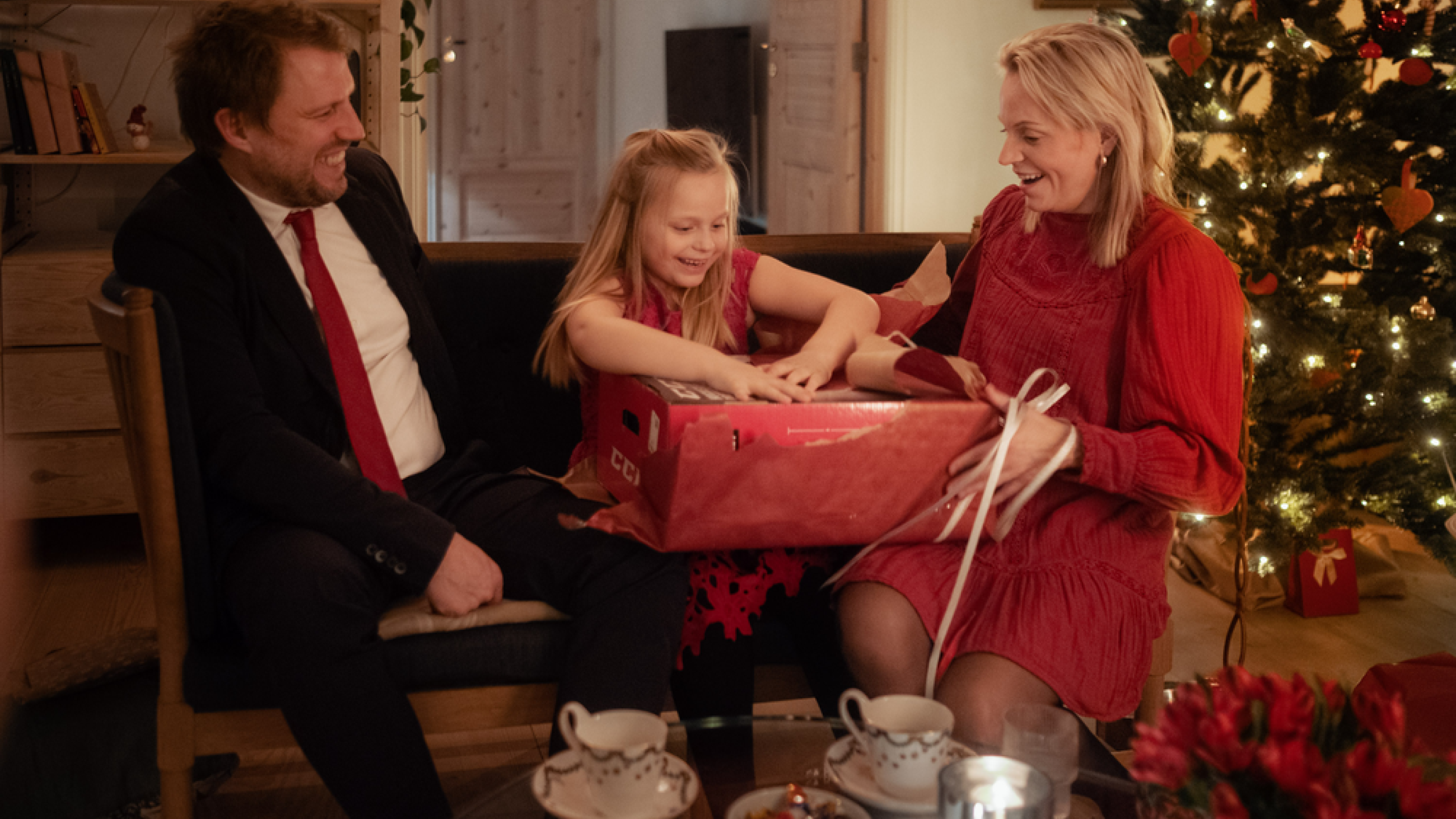 Far, mor og barn sitter i en sofa og jenta pakker opp en gave i rødt innpakningspapir.